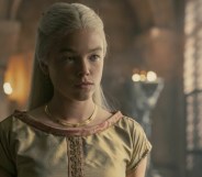 Photo of Rhaenyra dressing in a traditional off-white smock, standing in a grand palace room