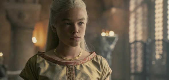 Photo of Rhaenyra dressing in a traditional off-white smock, standing in a grand palace room