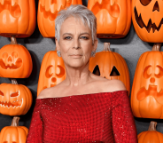 Jamie Lee Curtis wears a red outfit as she stands in front of a wall filled with orange pumpkin items shaped to look like jack-o-lanterns