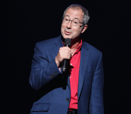 Ben Elton wears a suit while speaking into a microphone on stage