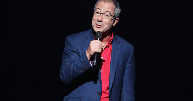 Ben Elton wears a suit while speaking into a microphone on stage