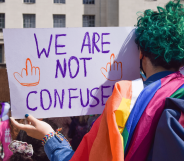 A person with green hair, who has their back turned to the camera, holds up a sign reading 'we are not confused' with an LGBTQ+ Pride flag draped along their shoulders