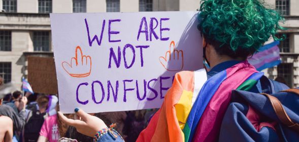 A person with green hair, who has their back turned to the camera, holds up a sign reading 'we are not confused' with an LGBTQ+ Pride flag draped along their shoulders