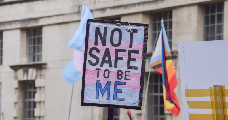 A person holds up a sign, designed in the colours of the trans Pride flag, that reads 'not safe to be me'
