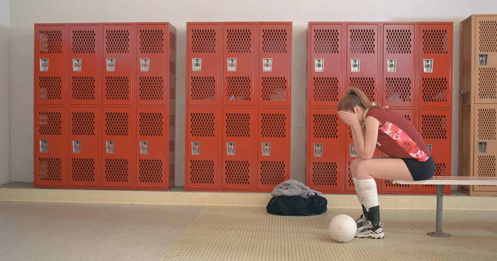 An image of a girl dressed in sports uniform seen sitting on a bench in a locker room holds her hands over her face like she is crying