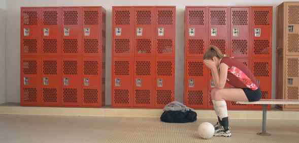 An image of a girl dressed in sports uniform seen sitting on a bench in a locker room holds her hands over her face like she is crying