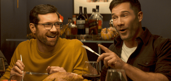 Billy Eichner and Luke Macfarlane at a dinner table, laughing with cutlery raised mid-gesture, in Bros