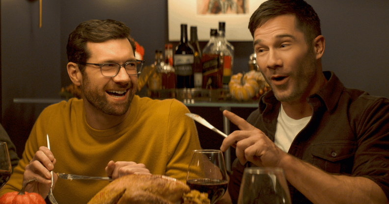Billy Eichner and Luke Macfarlane at a dinner table, laughing with cutlery raised mid-gesture, in Bros