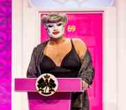 Drag Race UK queen Danny Beard standing at a pink podium in front of a pink front door