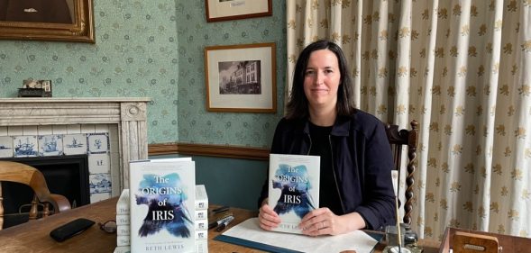 Author Beth Lewis wearing a black top and jacket sits behind a desk holding a copy of her book The Origins of Iris.