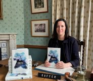 Author Beth Lewis wearing a black top and jacket sits behind a desk holding a copy of her book The Origins of Iris.