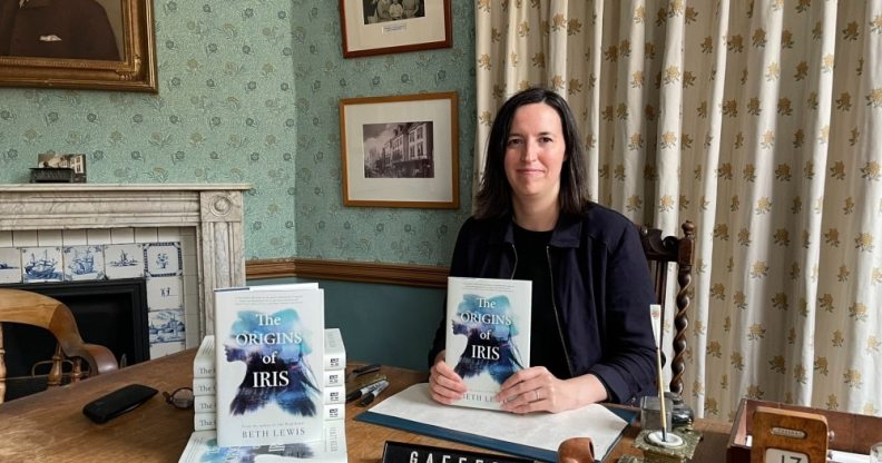 Author Beth Lewis wearing a black top and jacket sits behind a desk holding a copy of her book The Origins of Iris.