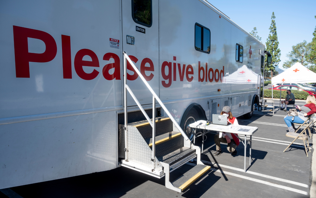 A blood donation van with the words "please give blood" is parked in a parking space, ready to accept donations.