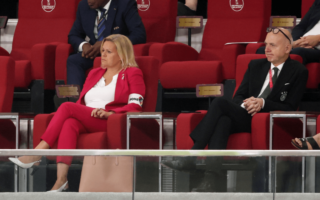 Seated in red cushioned seats at the Qatar World Cup stadium, Nancy Faeser is pictured in a red suit wearing the OneLove LGBTQ+ armband.