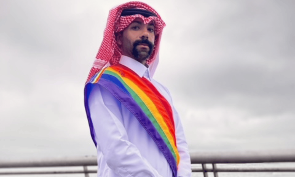 Dr Nas Mohamed wears Qatari attire with a sash in the colours of the LGBTQ+ Pride flag