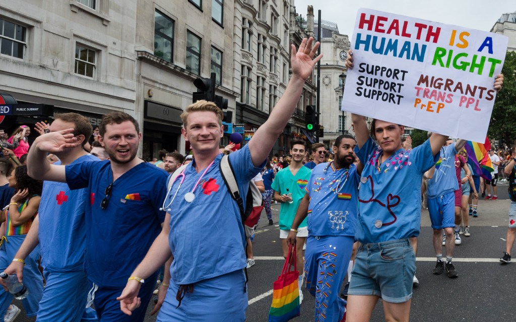 A group of NHS England doctors march during a Pride parade. One is wearing a sign that reads: "HEALTH IS A HUMAN RIGHT. Support Migrants, Support Trans PPL, Support PrEP"