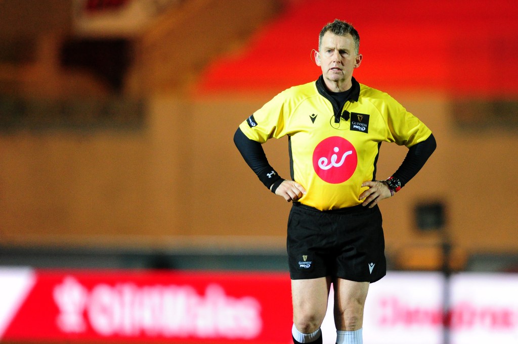 Nigel Owens standing with his hands on his hips during a rugby match wearing a yellow shirt and black trunks.