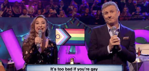 Screenshot of The Last Leg featuring actor Jess Robinson and presenter Adam Hills singing into microphones with a caption underneath saying "It's too bad i f you're gay"
