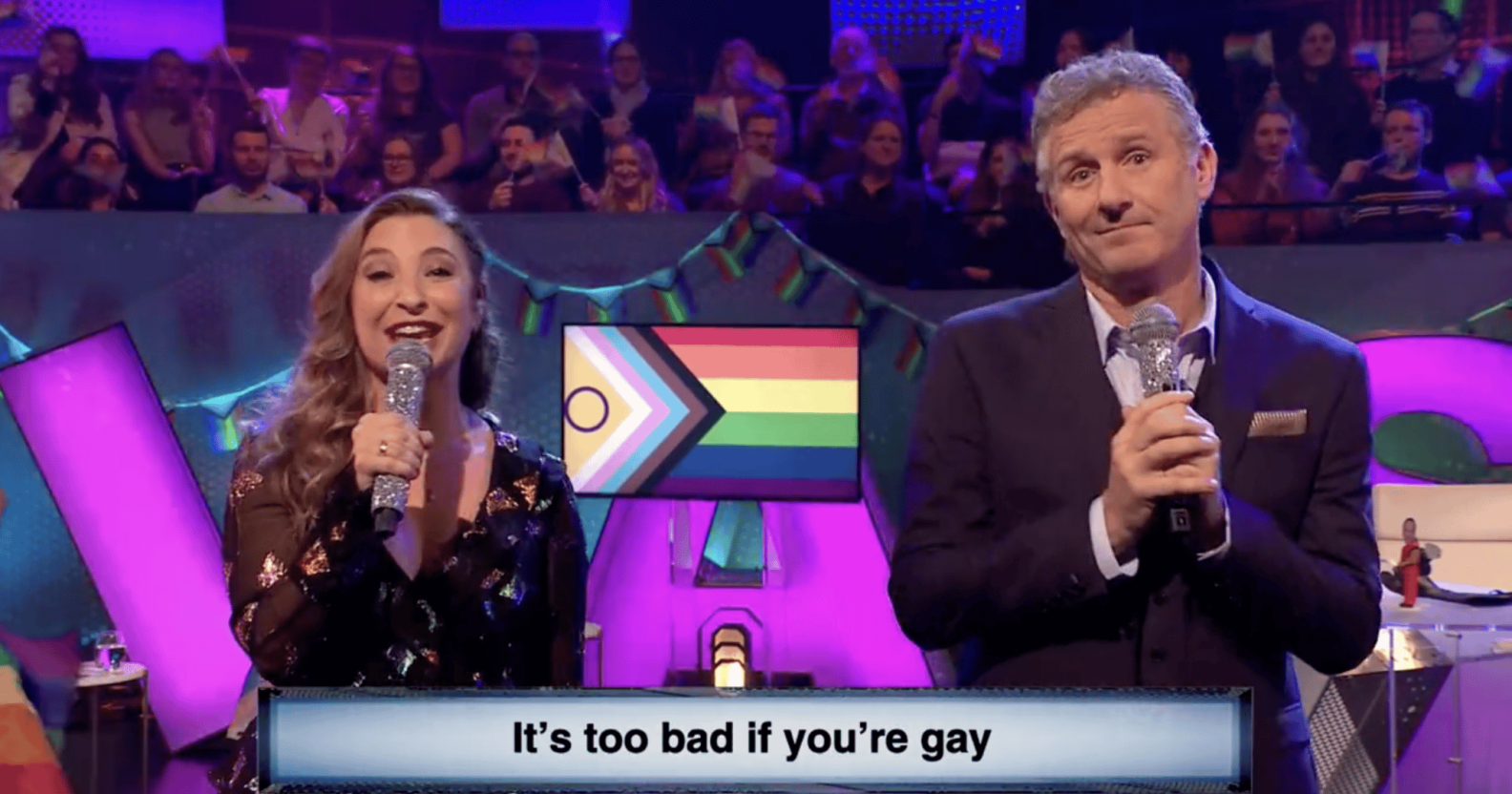 Screenshot of The Last Leg featuring actor Jess Robinson and presenter Adam Hills singing into microphones with a caption underneath saying "It's too bad i f you're gay"