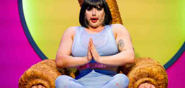 Drag queen Danny Beard in a bobbed wig and yoga gear, sitting cross-legged in a Big Brother-style 'Diary Room chair'