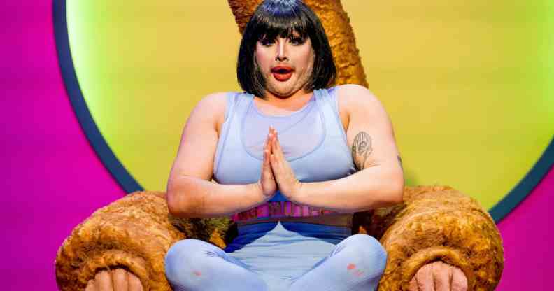 Drag queen Danny Beard in a bobbed wig and yoga gear, sitting cross-legged in a Big Brother-style 'Diary Room chair'