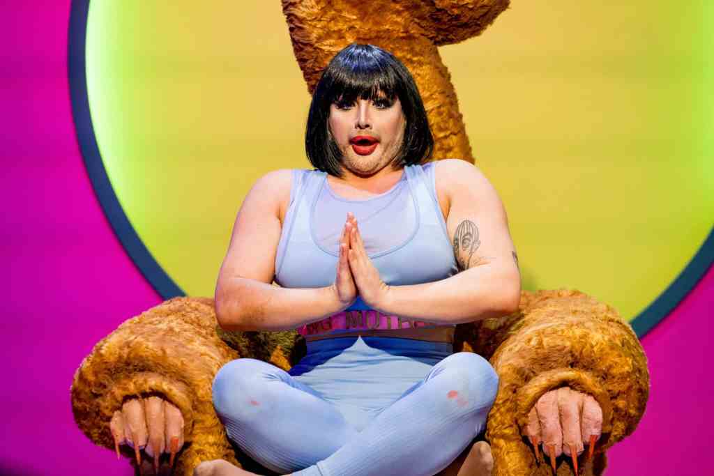 Drag queen Danny Beard in a bobbed wig and yoga gear, sitting cross-legged in a Big Brother-style 'Diary Room chair'