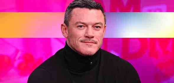Actor Luke Evans wearing a black top sits in front of a pink background