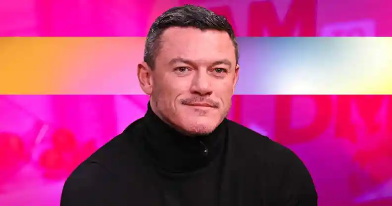 Actor Luke Evans wearing a black top sits in front of a pink background