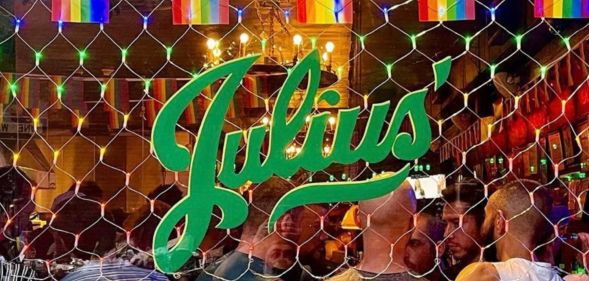 A photo showing an exterior shot of New York gay bar Julius' - showing Pride flags across the top, the green Julius' logo and people on the inside of the club