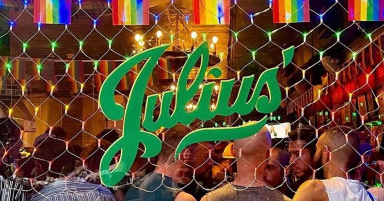 A photo showing an exterior shot of New York gay bar Julius' - showing Pride flags across the top, the green Julius' logo and people on the inside of the club