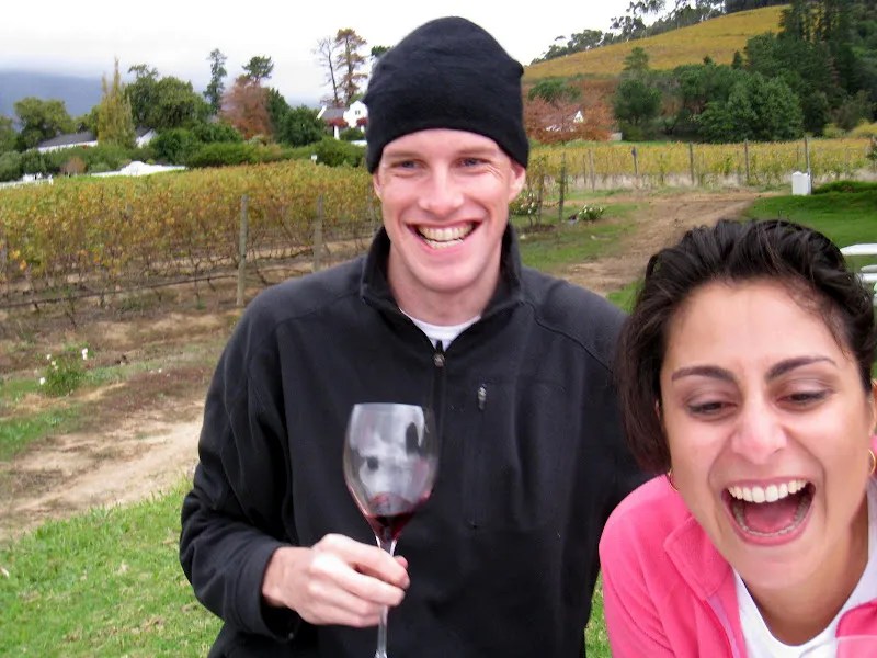 A photo of late journalist Grant Wahl holding a glass of wine and wearing a black jacket and wooly hat with his wife Céline Gounder who is wearing a pink top.