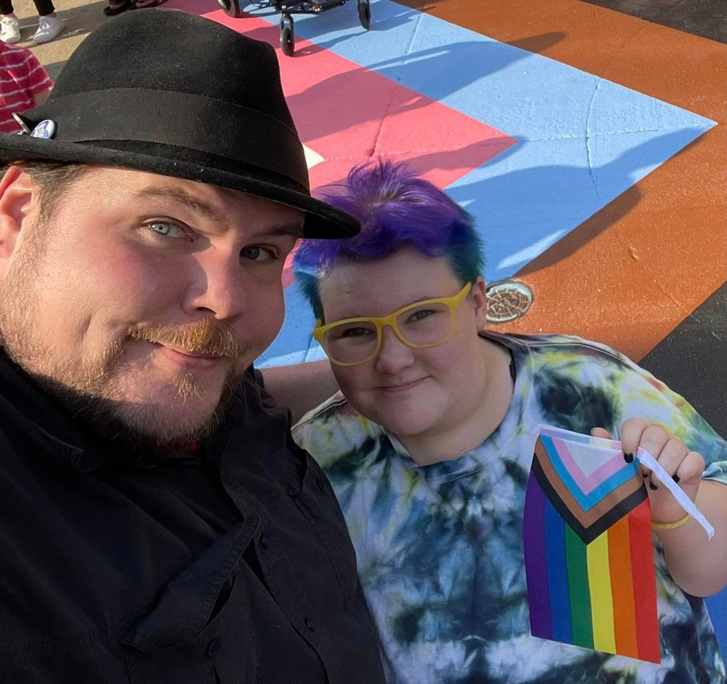 A photo of chef Dave Heide dressed in black and wearing a black hat with his arm round his child Ollie who's wearing a tie-dye t-shirt, yellow glasses and is holding a Pride flag