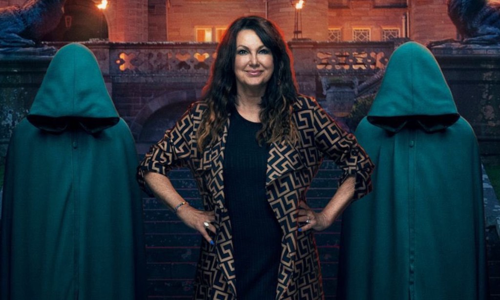 A promo image from BBC's The Traitors showing contestant Amanda Lovett dressed in black with a black and gold jacket standing in front of a gothic-looking building with two figures standing either side of her wearing green hooded cloaks and having their heads bowed so you can't see their faces