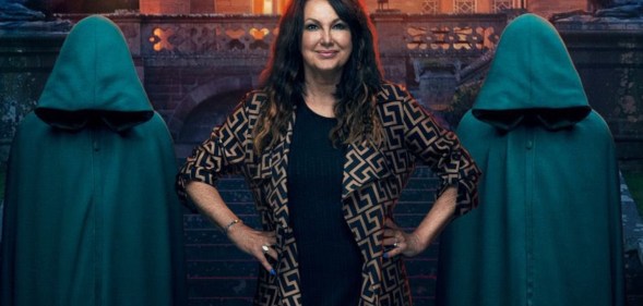 A promo image from BBC's The Traitors showing contestant Amanda Lovett dressed in black with a black and gold jacket standing in front of a gothic-looking building with two figures standing either side of her wearing green hooded cloaks and having their heads bowed so you can't see their faces