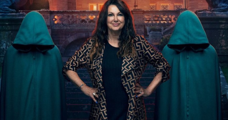 A promo image from BBC's The Traitors showing contestant Amanda Lovett dressed in black with a black and gold jacket standing in front of a gothic-looking building with two figures standing either side of her wearing green hooded cloaks and having their heads bowed so you can't see their faces