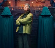 A promo photo from BBC's The Traitors showing contestant Andrea dressed in a dark green top and light green cardigan with her arms crossed standing on some steps to a gothic-looking building with two people standing either side of her wearing hooded cloaks and their heads bowed so as to hide their faces. (BBC/Studio Lambert Associates/Mark Mainz)