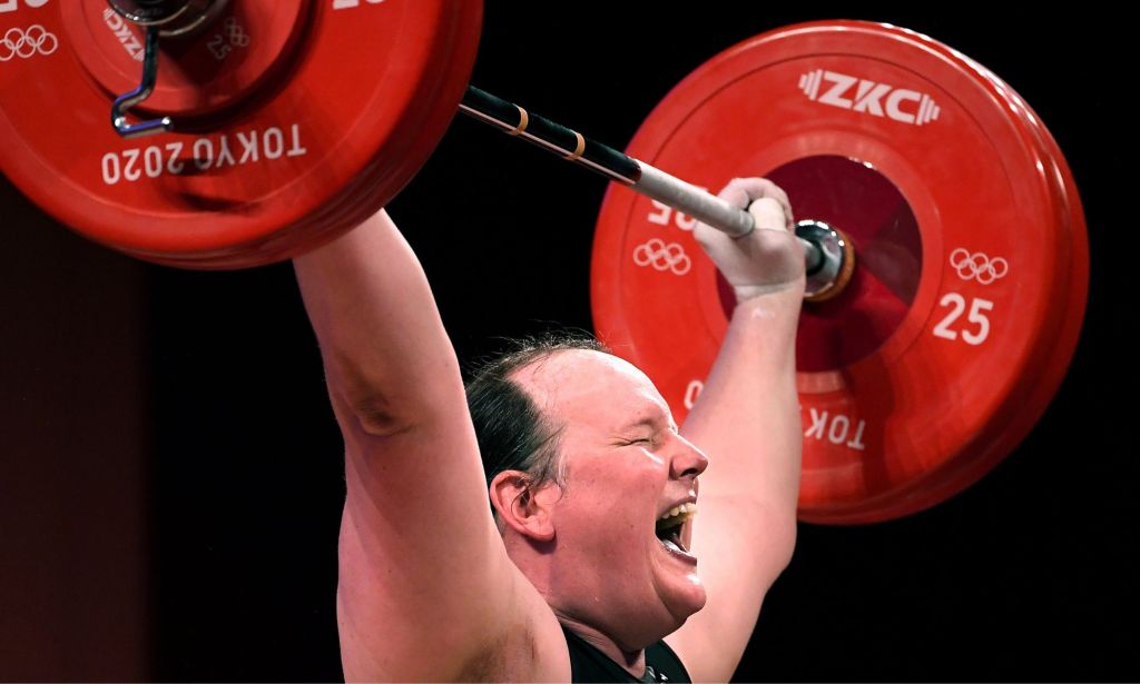 A photo of New Zealand trans athlete weightlifter Laurel Hubbard showing signs of physical strain as she lifts a dumbbell weight over her head during the Olympics.