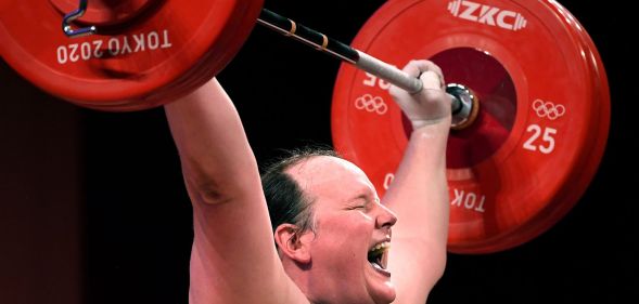 A photo of New Zealand trans athlete weightlifter Laurel Hubbard showing signs of physical strain as she lifts a dumbbell weight over her head during the Olympics.