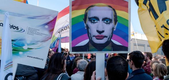 Several protestors march down a street, as a member of the public holds up a picture of Vladimir Putin, edited so that he is wearing makeup.