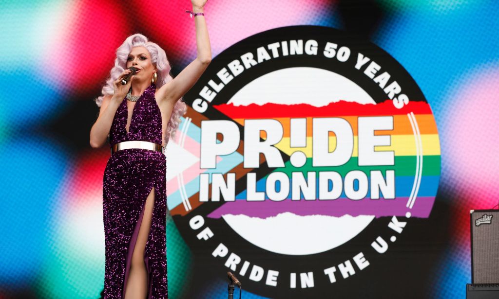 A drag queenw earing pink hair speaks on stage, which a giant Prirde in London sticker graphic behind her.
