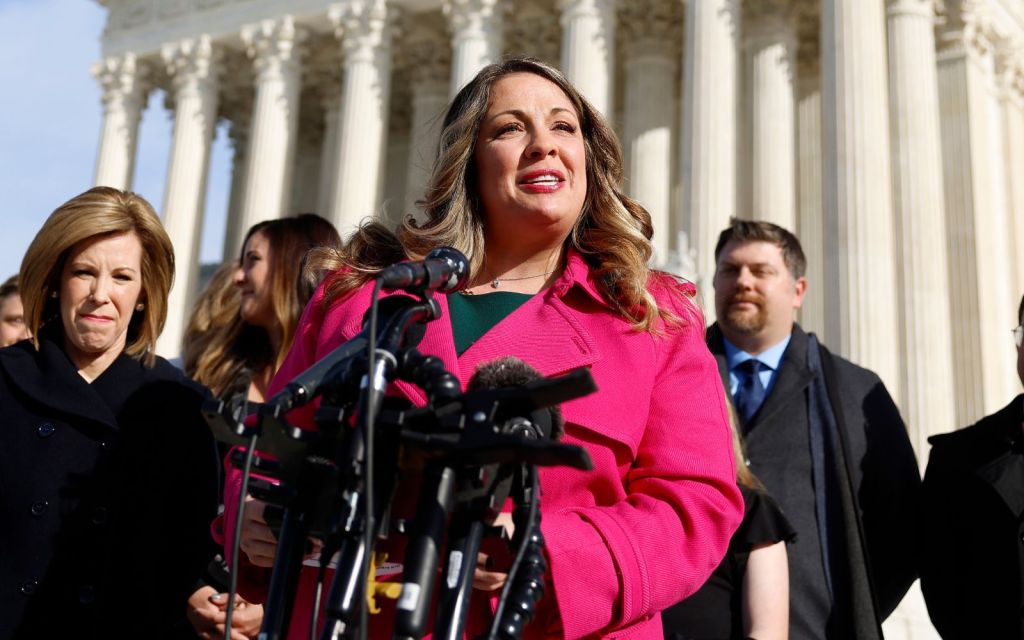 Lorie Smith, in a pink jacket, speaks outside the Supreme Court, with an array of microphones in front of her.
