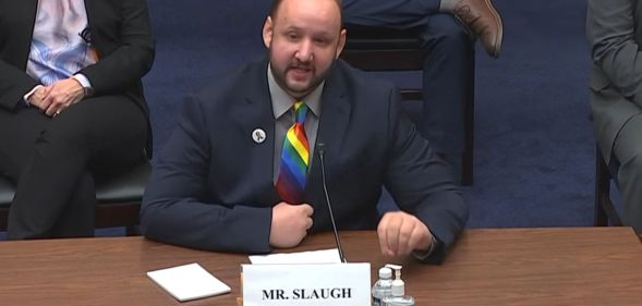 James Slaugh, wearing a rainbow tie and black blazer, speaks to a committe while several people sit behind him during a scheduled meeting.