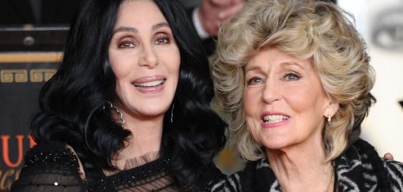 Cher, wearing a black dress, holds her mother, who is wearing a grey striped suit.