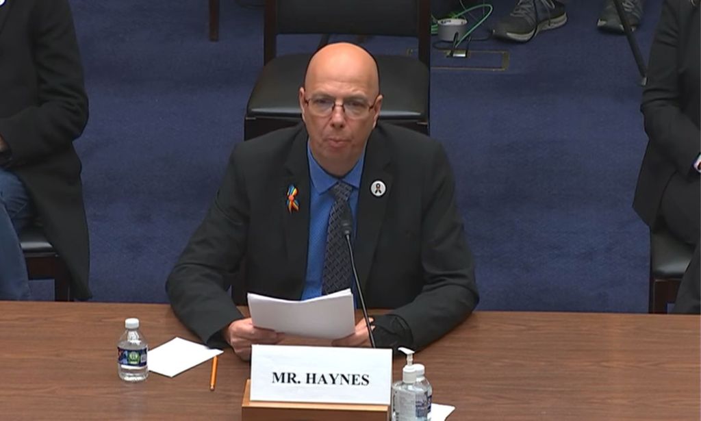 Matthew Haynes speaks at a wooden table during a testimony for the anti-LGBTQ+ hate speech committee, sat either side of fellow witnesses and holding a piece of paper.