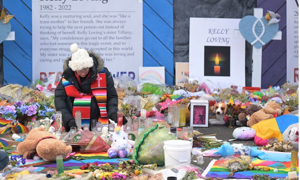 An individual in a coat, scarf, and white hat sits amid the various tributes to those who died in the Club Q shooting.