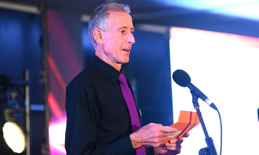 Peter Tatchell, in a purple tie, speaks to an audience through a microphone stand while holding a card.