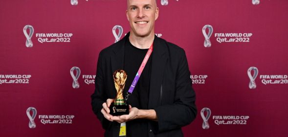Grant Wahl stands infront of a red back drop with the Qatar World Cup logo written across it, while holding a miniature replica of the World Cup trophy, wearing a black blazer.
