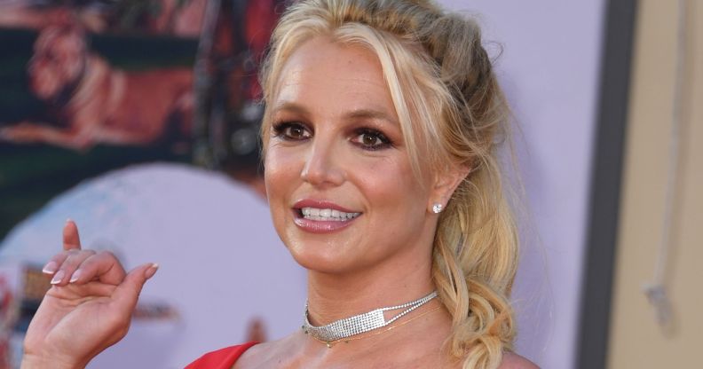 Britney spears, smiling and wearing a red dress, waves at onlookers as she walks past.