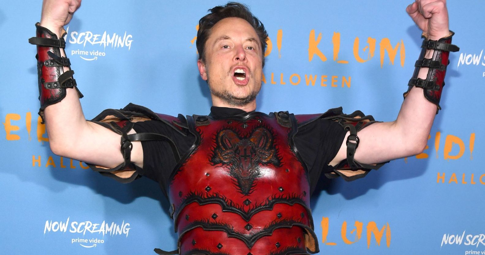 Elon Musk, wearing a red suit of armour, raises his arms and screams.