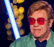 Elton John, wearing red glasses, biting his lip and looking to the right.